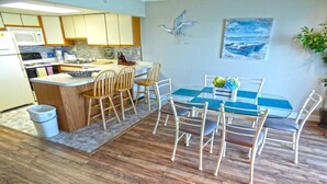 A modern kitchen with wooden cabinets, a microwave, and a coffee maker. There is a dining area with a glass table and chairs. - Harbour Island 306l 2 Bedroom Condo by RedAwning (Ocean City)
