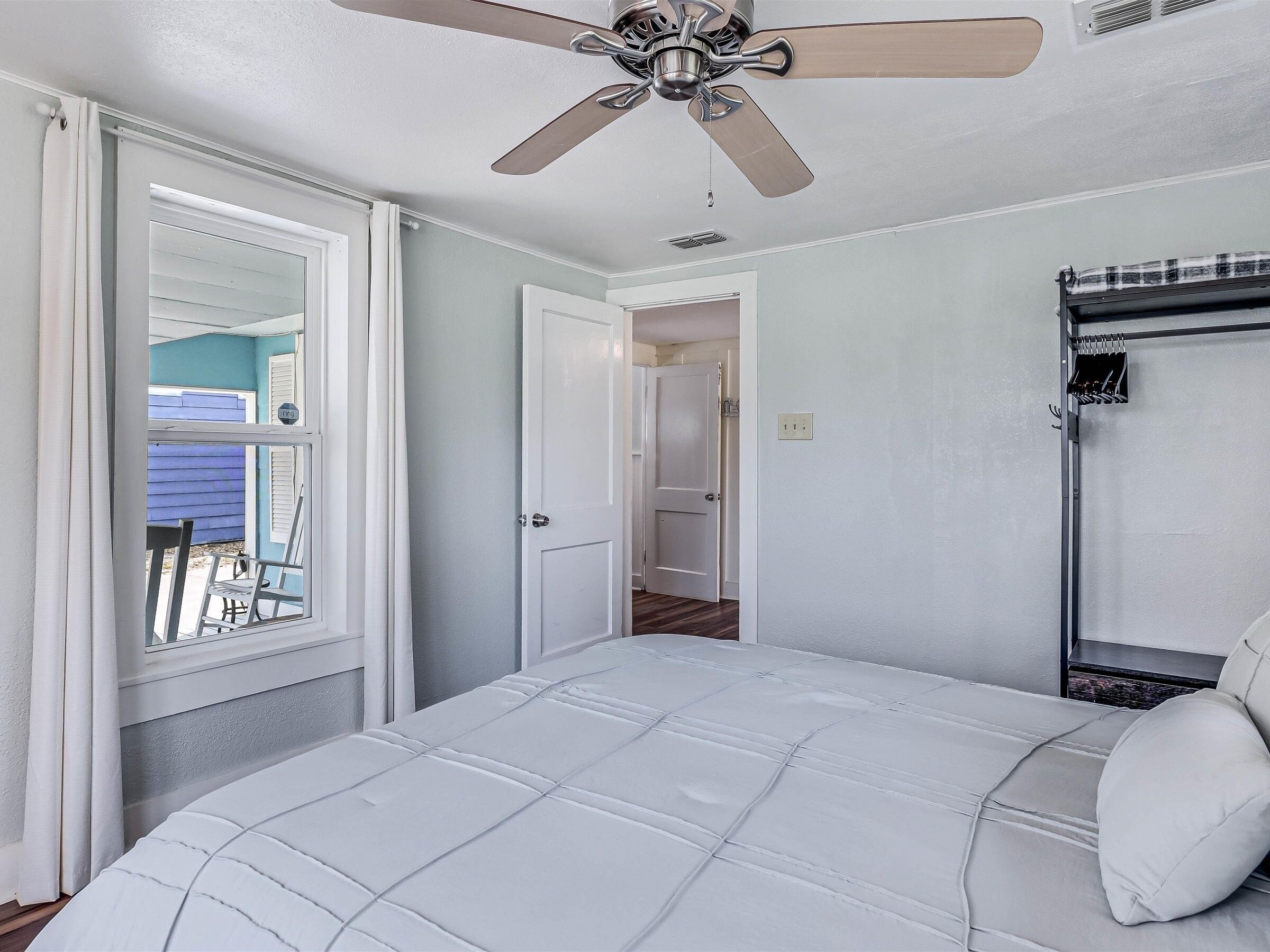 Un dormitorio con una cama, ventilador de techo, ventana con cortinas y puerta que da a otra habitación.