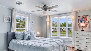 A bedroom with a bed, a ceiling fan, a window with a view of the outdoors, and a painting on the wall.