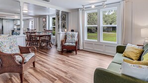 A spacious living area with a dining table, wicker chairs, a sofa, and large windows offering a view of the outdoors.