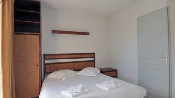 A neatly made bed with white linens and towels, a wooden headboard, a small bedside table, and a wardrobe with a wicker panel.