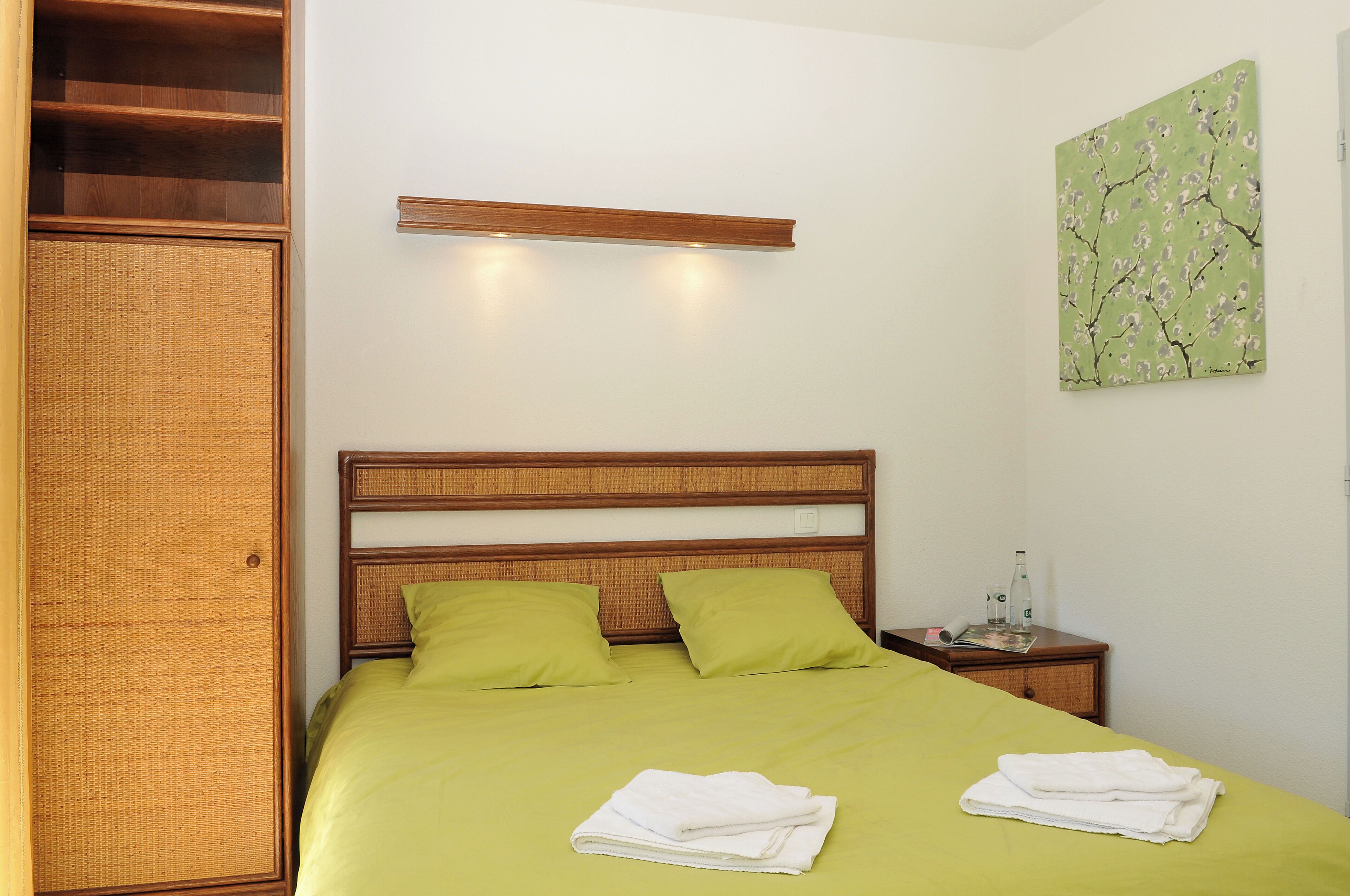A neatly made bed with green bedding and pillows, a wooden headboard, a nightstand with a bottle and glasses, and a wall-mounted shelf above the bed.
