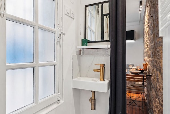 Ein Badezimmer mit einem weißen Waschbecken, einem goldenen Wasserhahn, einem Spiegel und einem Fenster.