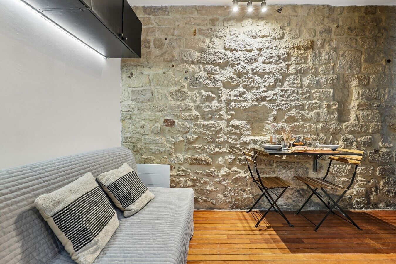 Une pièce avec un mur en pierre, une table à manger en bois avec des chaises, un canapé gris avec des coussins et un plancher en bois.