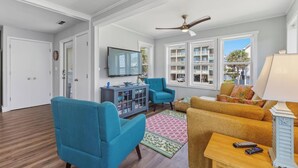 A living room with a ceiling fan, a TV, a blue armchair, a yellow sofa, and a window with a view of a building. - 🌴 Surf Haus: Fenced Yard | 1 Block to the Beach | Screened-In Porch 🌴 (Carolina Beach)