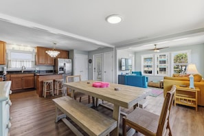 A modern kitchen with wooden cabinets, a stainless steel refrigerator, and a dining area with a wooden table and benches. - 🌴 Surf Haus: Fenced Yard | 1 Block to the Beach | Screened-In Porch 🌴 (Carolina Beach)
