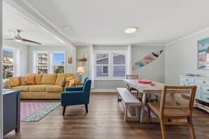 A living room with a sofa, a blue armchair, a wooden dining table, and a bench. - 🌴 Surf Haus: Fenced Yard | 1 Block to the Beach | Screened-In Porch 🌴 (Carolina Beach)