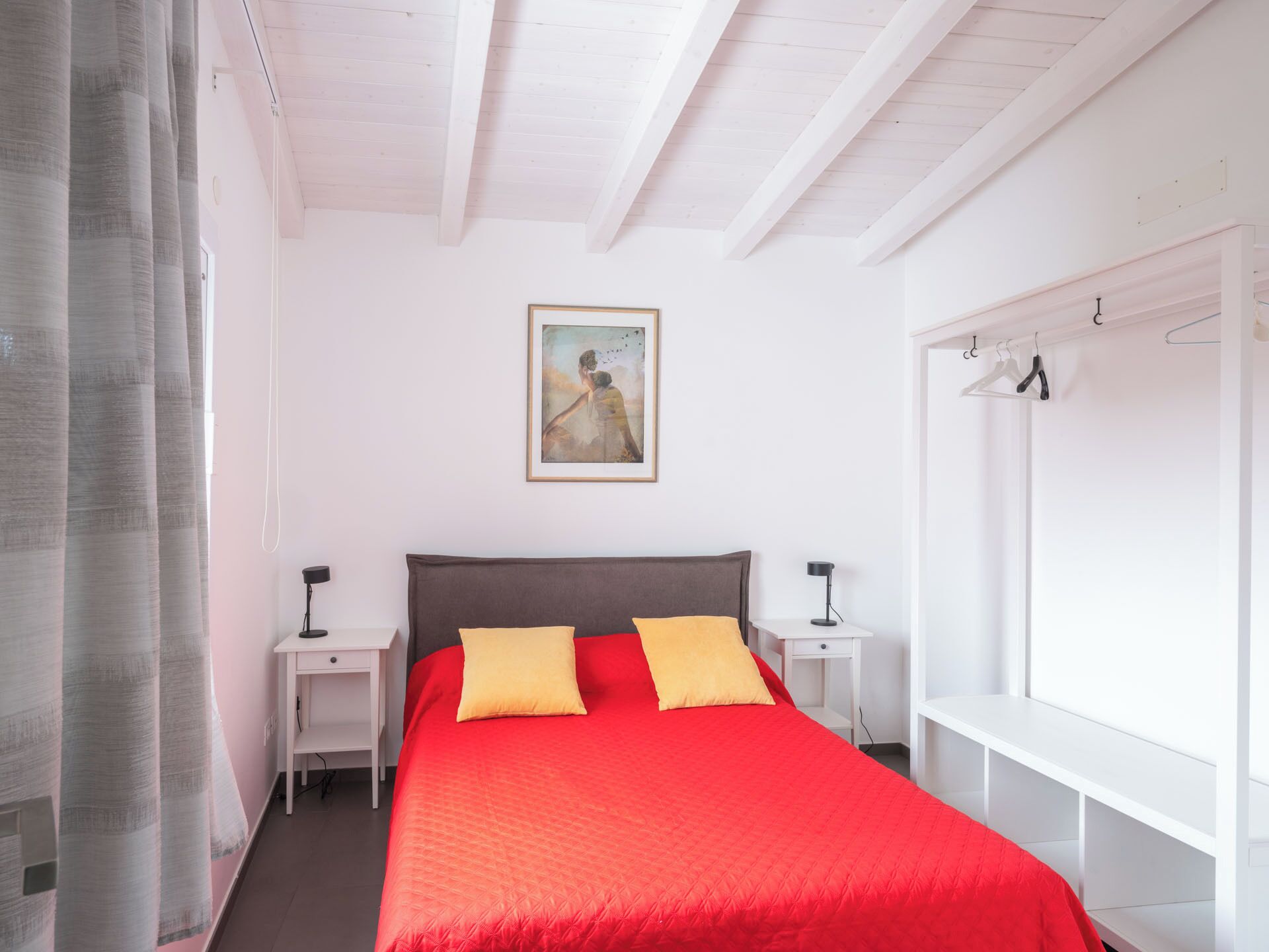A bedroom with a large bed, two bedside tables, a nightstand, a painting on the wall, and a window with a curtain.