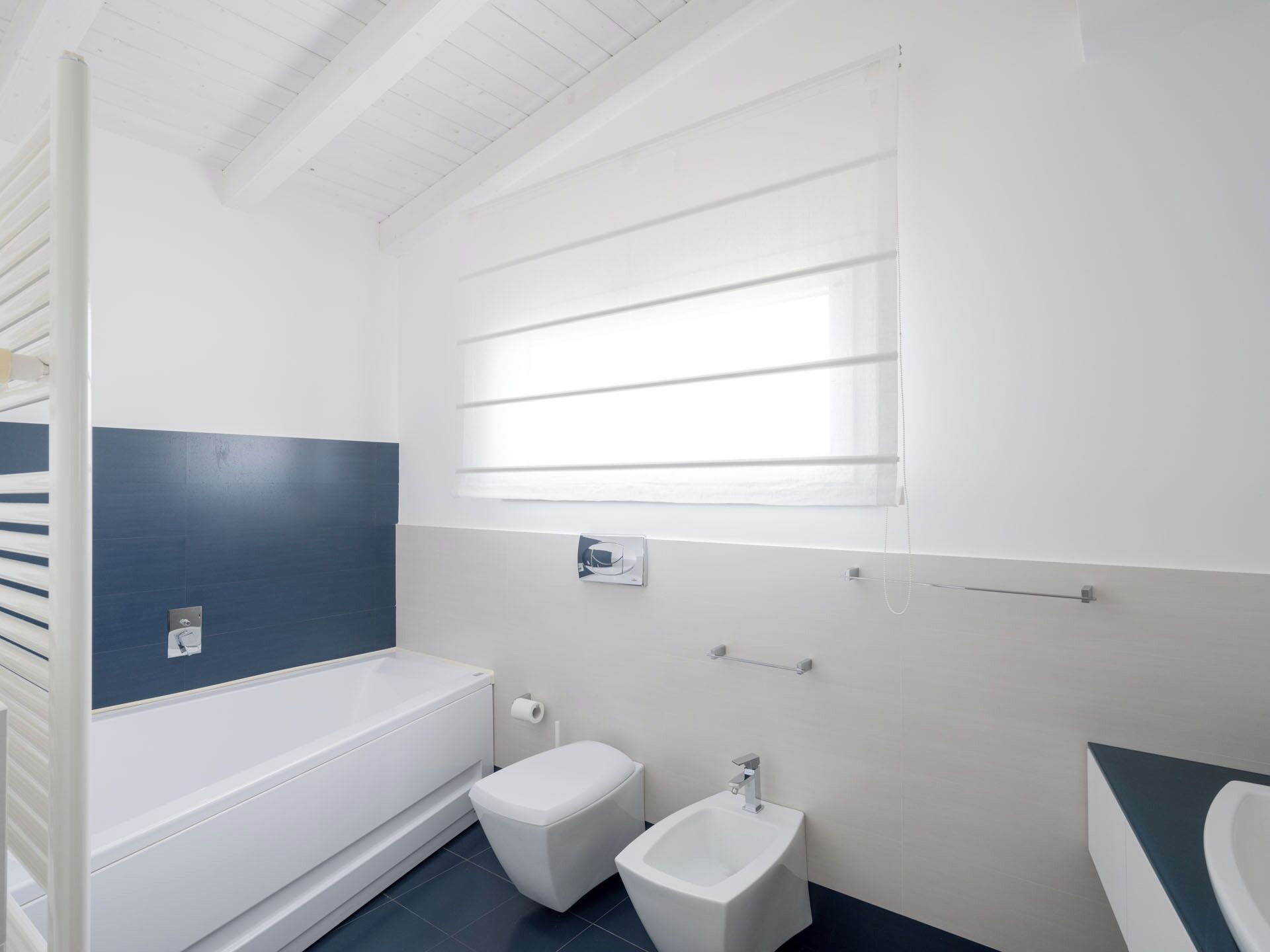 Een moderne badkamer met een bad, toilet, bidet en een raam met zonwering.