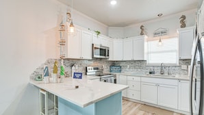 A modern kitchen with white cabinets, a central island, stainless steel appliances, and decorative elements like a hanging light fixture and a sign reading "Beach Rules". - Serene Surfside Beach Retreat w/ Ocean Views! (Freeport)