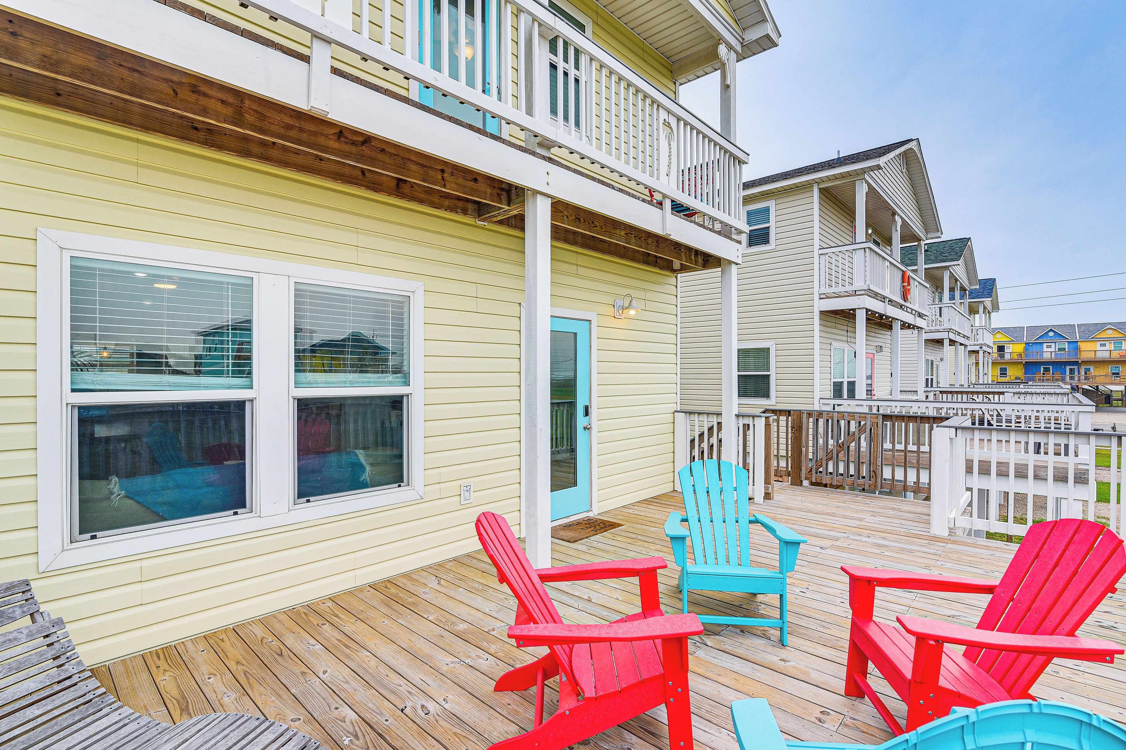 A wooden deck with red and blue Adirondack chairs, a white door, and a yellow building with a balcony.