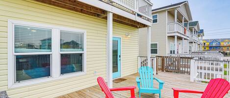 A wooden deck with red and blue Adirondack chairs, a white door, and a yellow building with a balcony.