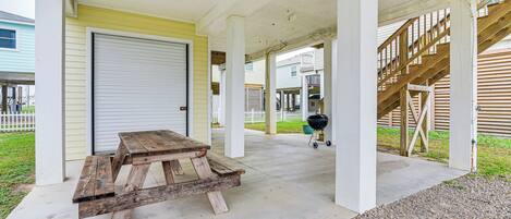 A covered patio area with a wooden picnic table, a barbecue grill, and a garage door.