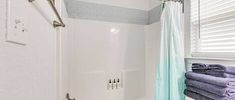 A shower area with a glass door, a towel rack with folded towels, and a window with blinds.