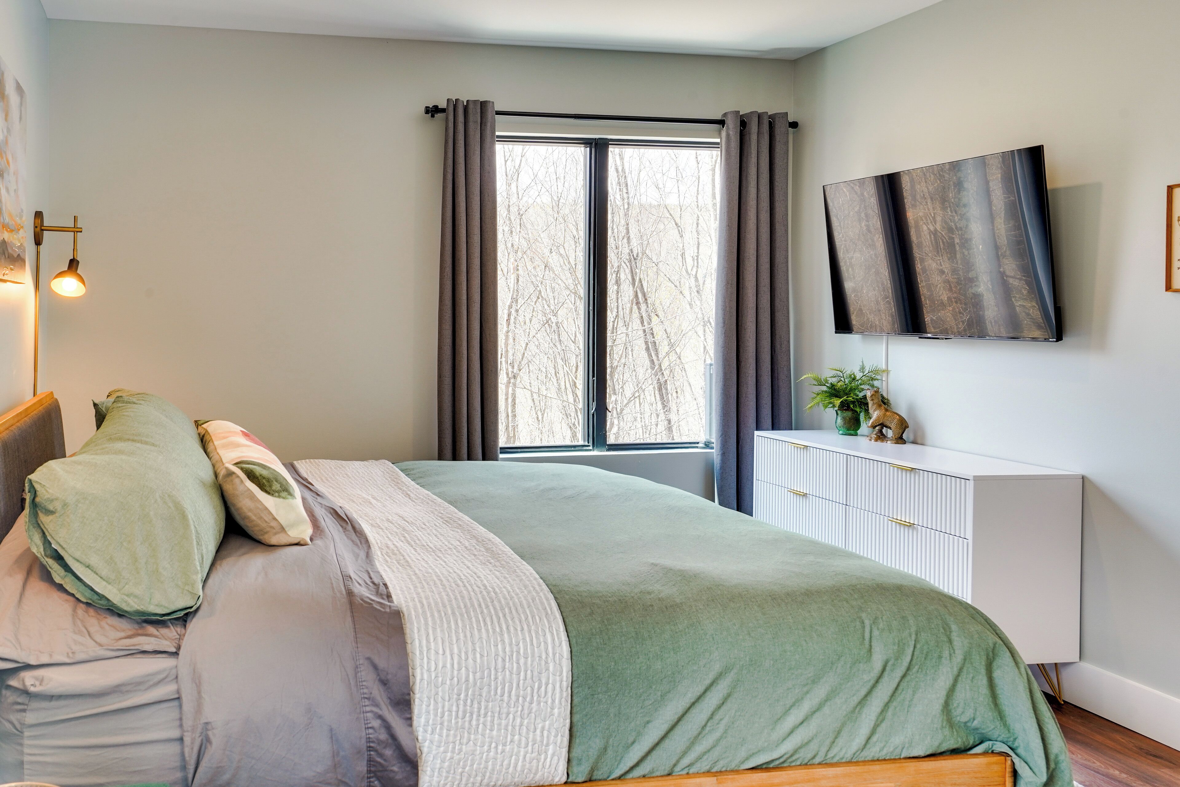 A bedroom with a bed, a TV mounted on the wall, a window with curtains, and a bedside plant.