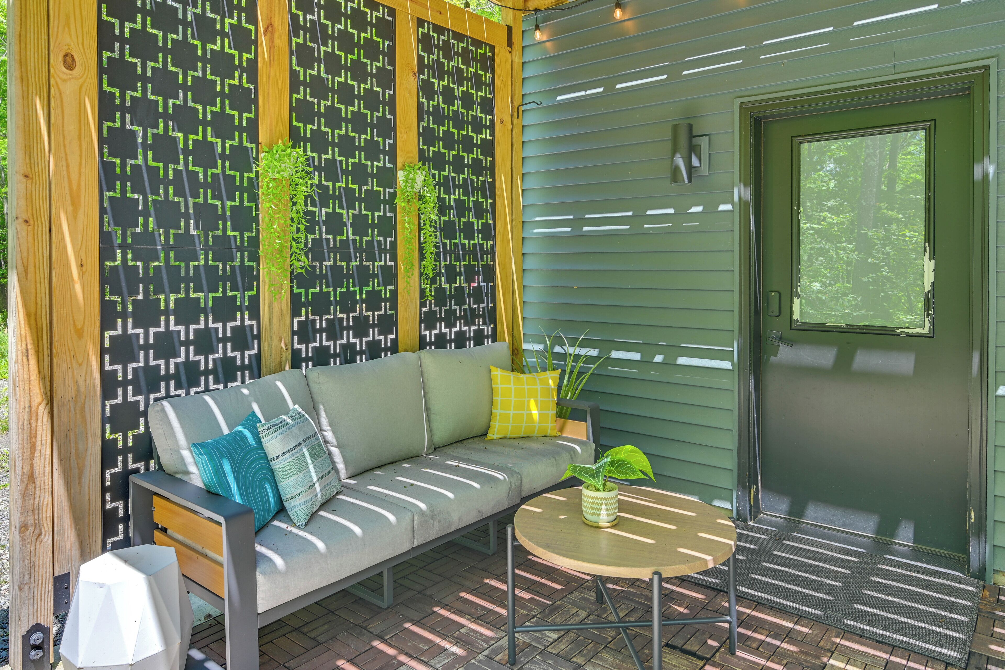 A covered outdoor seating area with a sofa, a round table, and a door leading to a green exterior.