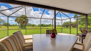 A conservatory with a large table and striped sofas, a vase with flowers, and a view of a green landscape with trees and a clear sky.