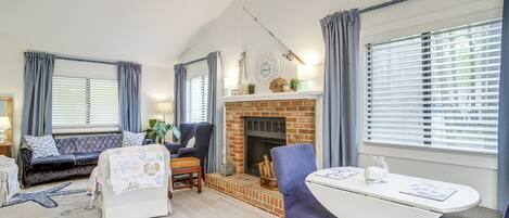 A cozy living room with a fireplace, a ceiling fan, and a dining area with blue chairs.