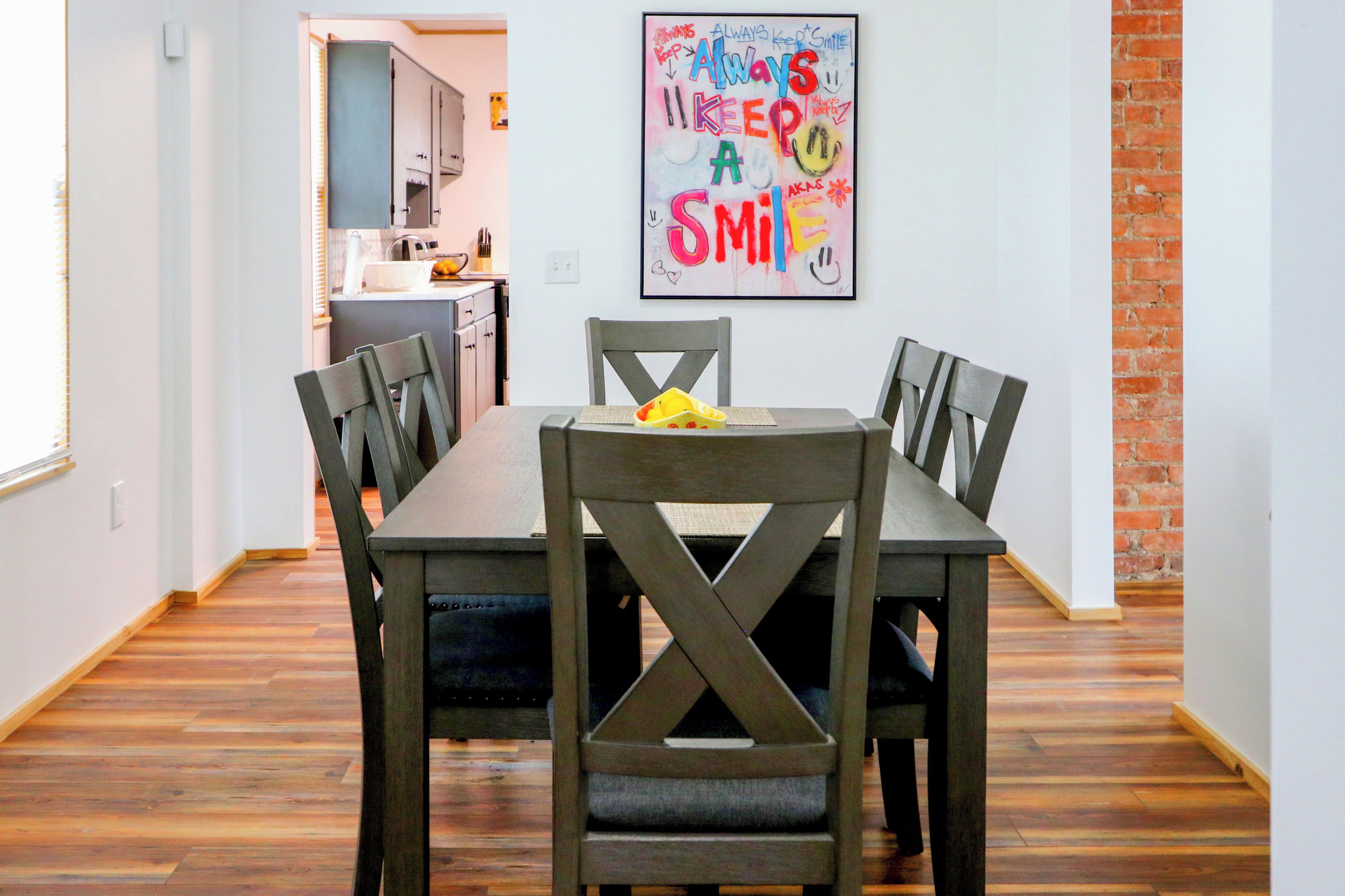 Espaço de refeições com mesa e cadeiras de madeira, cozinha ao fundo e uma obra de arte colorida na parede.