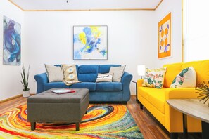 A living room with a blue sofa, a yellow sofa, and a grey ottoman. There are abstract paintings on the walls and a colorful rug on the floor. - Charming Home - 2 Mi to Niagara Falls State Park! (Niagara Falls)