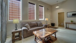 A living room with a sofa, coffee table, and two lamps. There are three windows with blinds, a door, and a small table with a lamp. - Riverfront Briceville Apt: Atv, Hike & More! (Briceville)