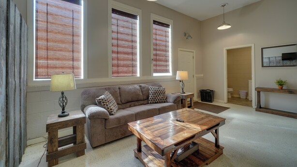 A living room with a sofa, coffee table, and two lamps. There are three windows with blinds, a door, and a small table with a lamp. - Riverfront Briceville Apt: Atv, Hike & More! (Briceville)