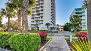 A modern hotel with multiple floors, a central courtyard with palm trees, and a well-maintained garden with pink flowers. - Ocean-view Balcony: Peaceful Forest Dunes Condo! (Myrtle Beach)