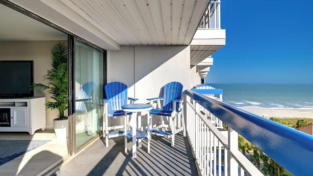 A balcony with blue chairs and a table, overlooking a beach. - Ocean-view Balcony: Peaceful Forest Dunes Condo! (Myrtle Beach)