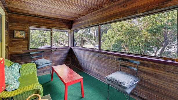 Une pièce aux murs en bois et aux grandes fenêtres, avec un canapé vert, une table rouge et un banc doté d’un coussin.