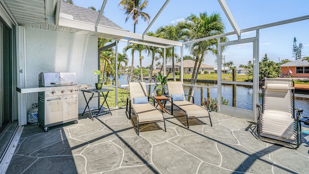 Un patio al aire libre con una parrilla, sillas de descanso y vistas a un lago y palmeras.