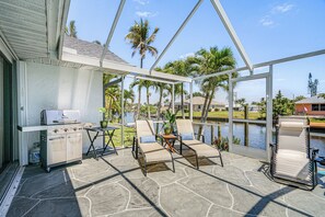 Une terrasse extérieure équipée d’un barbecue, de chaises longues et offrant une vue sur un lac et des palmiers.