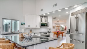 Una cocina moderna con armarios blancos, una isla central con sillas, un refrigerador de acero inoxidable y un área de comedor con mesa de billar.