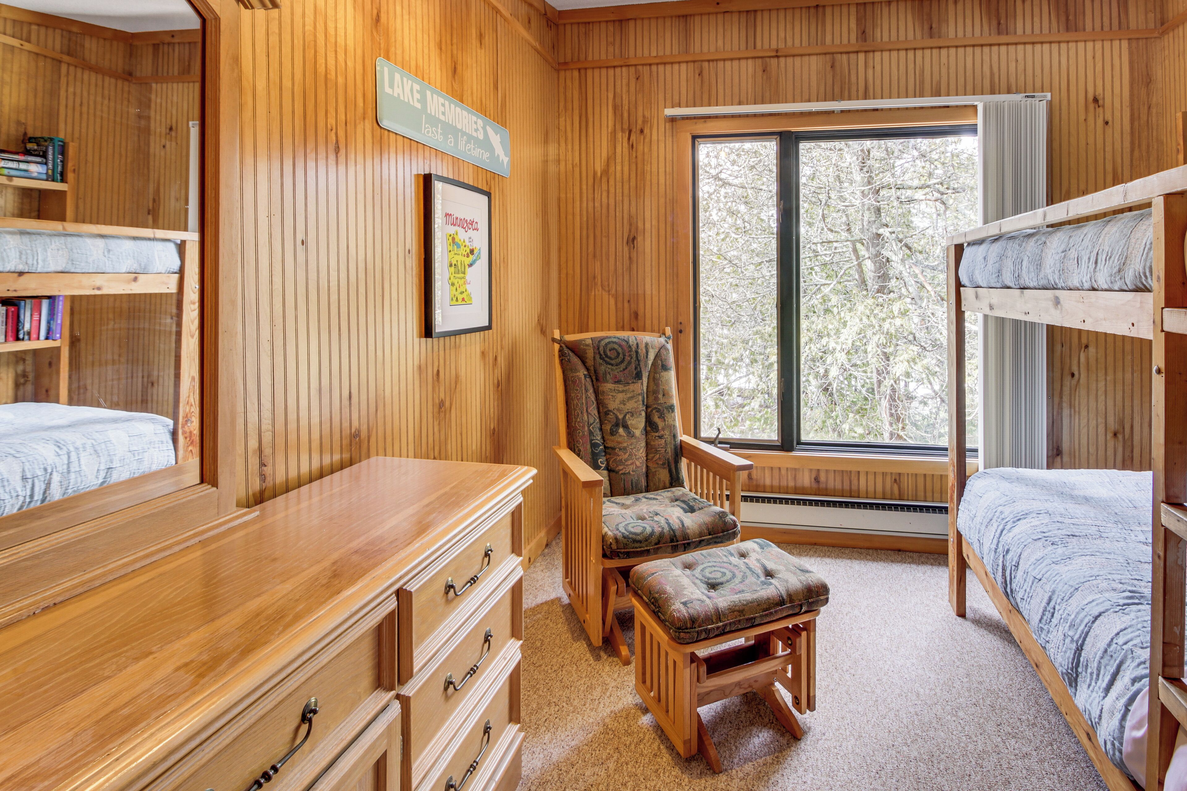 A cozy cabin-style room with wooden walls, a large window, bunk beds, a wooden dresser, and a chair with a cushion.