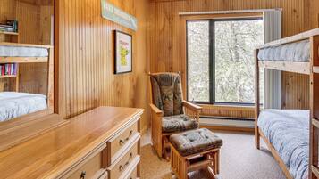 A cozy cabin-style room with wooden walls, a large window, bunk beds, a wooden dresser, and a chair with a cushion.