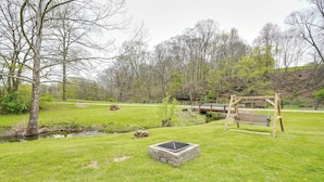 A grassy outdoor area with a stone fire pit, a wooden swing set, and a small bridge over a stream. - Pet-friendly Zanesville Haven w/ Game Room! (Zanesville)