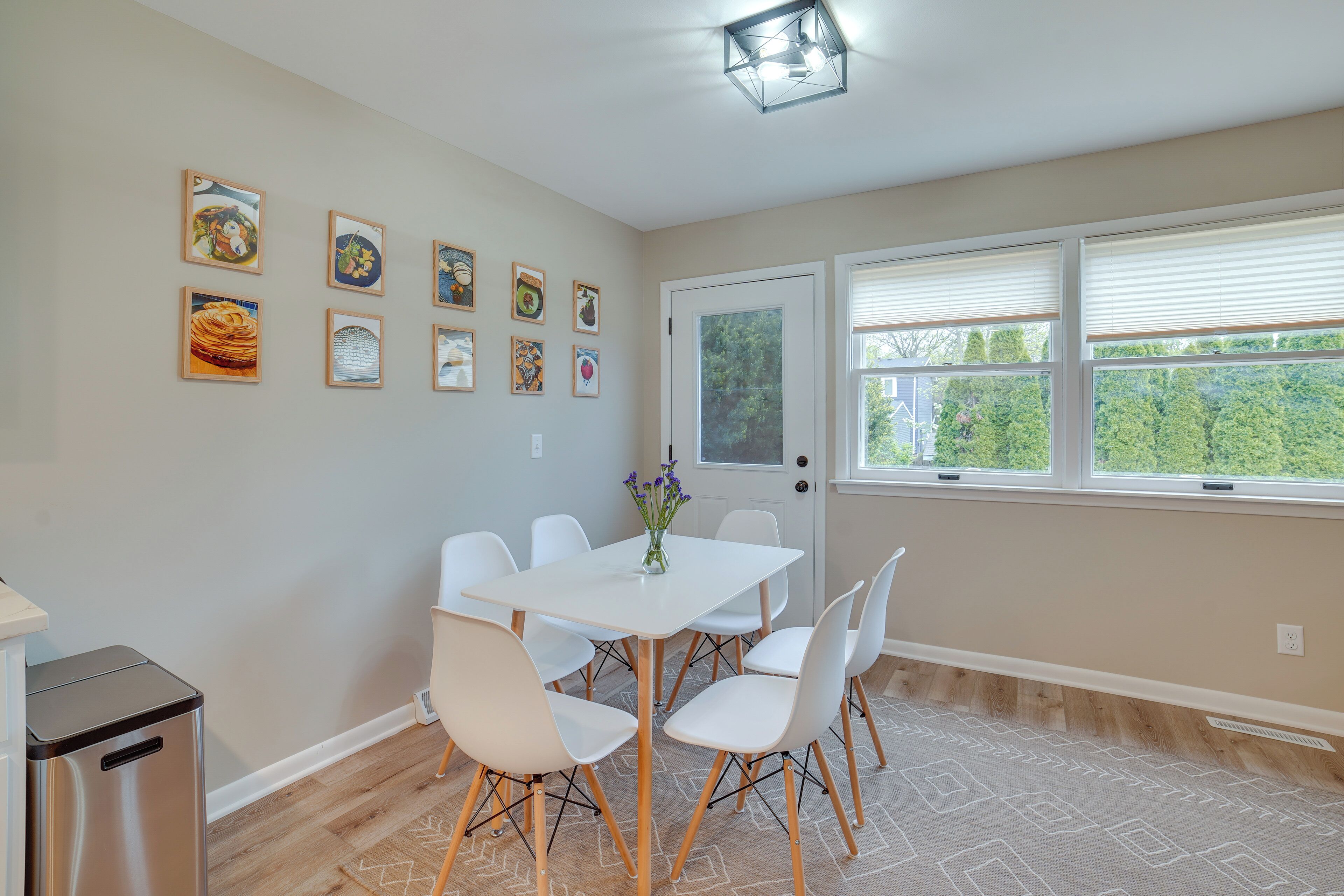 A dining area with a white table and chairs, framed artwork on the wall, and a window with blinds.