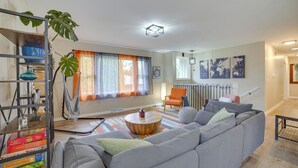 Una sala moderna con un sofá gris, una mesa redonda de madera y una alfombra colorida. Se observa una estantería con juegos de mesa y una planta grande.