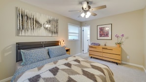 Un dormitorio con una cama, ventilador de techo, cómoda y un cuadro en la pared.