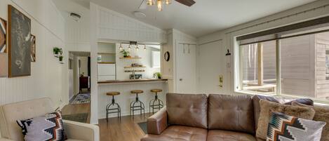 A modern living room with a brown leather sofa, a white armchair, and a patterned rug. There is a ceiling fan, a kitchen area in the background, and a large window providing natural light.
