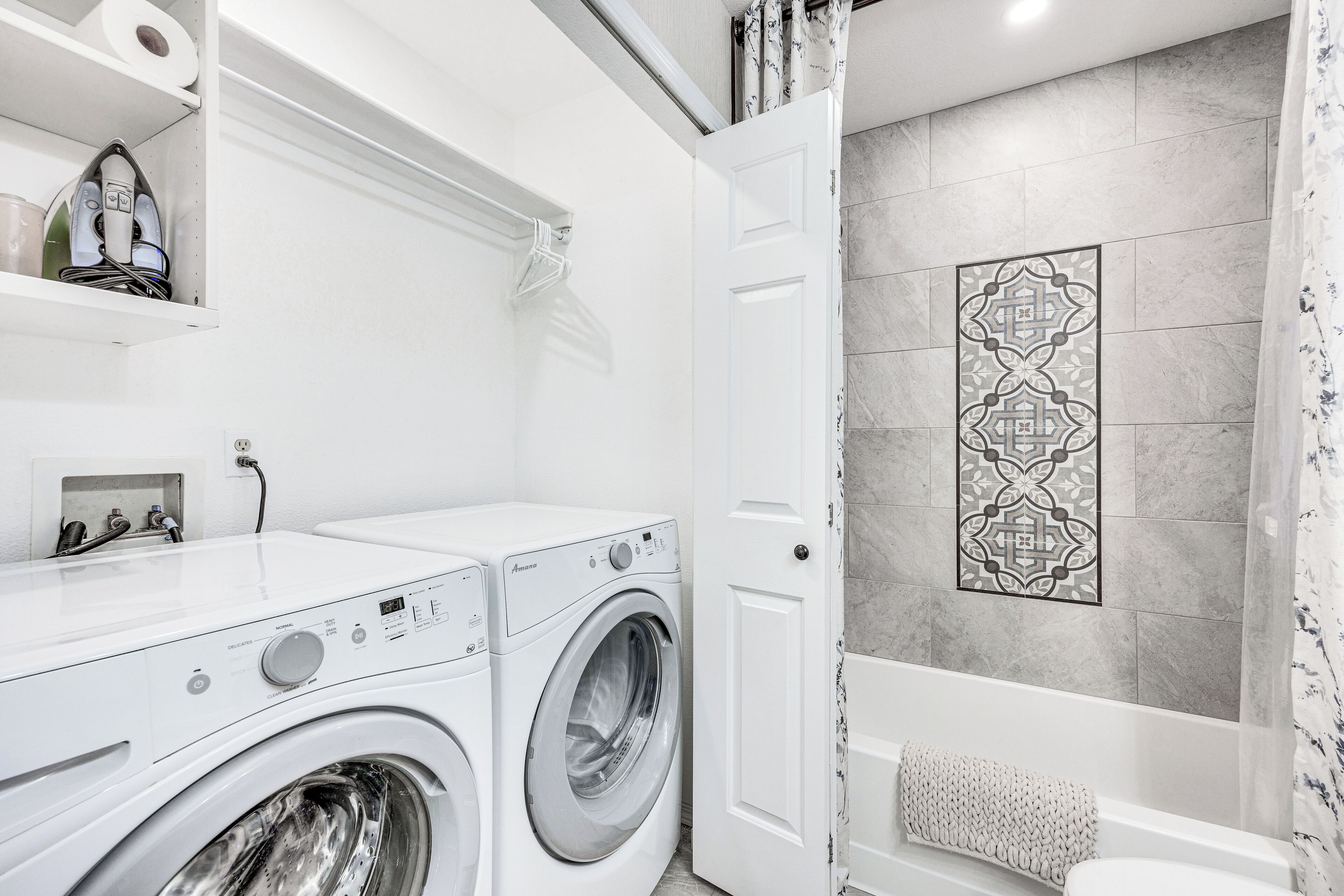 A modern laundry room with a front-loading washing machine and dryer, a shelf with cleaning supplies, and a decorative wall piece above the bathtub.