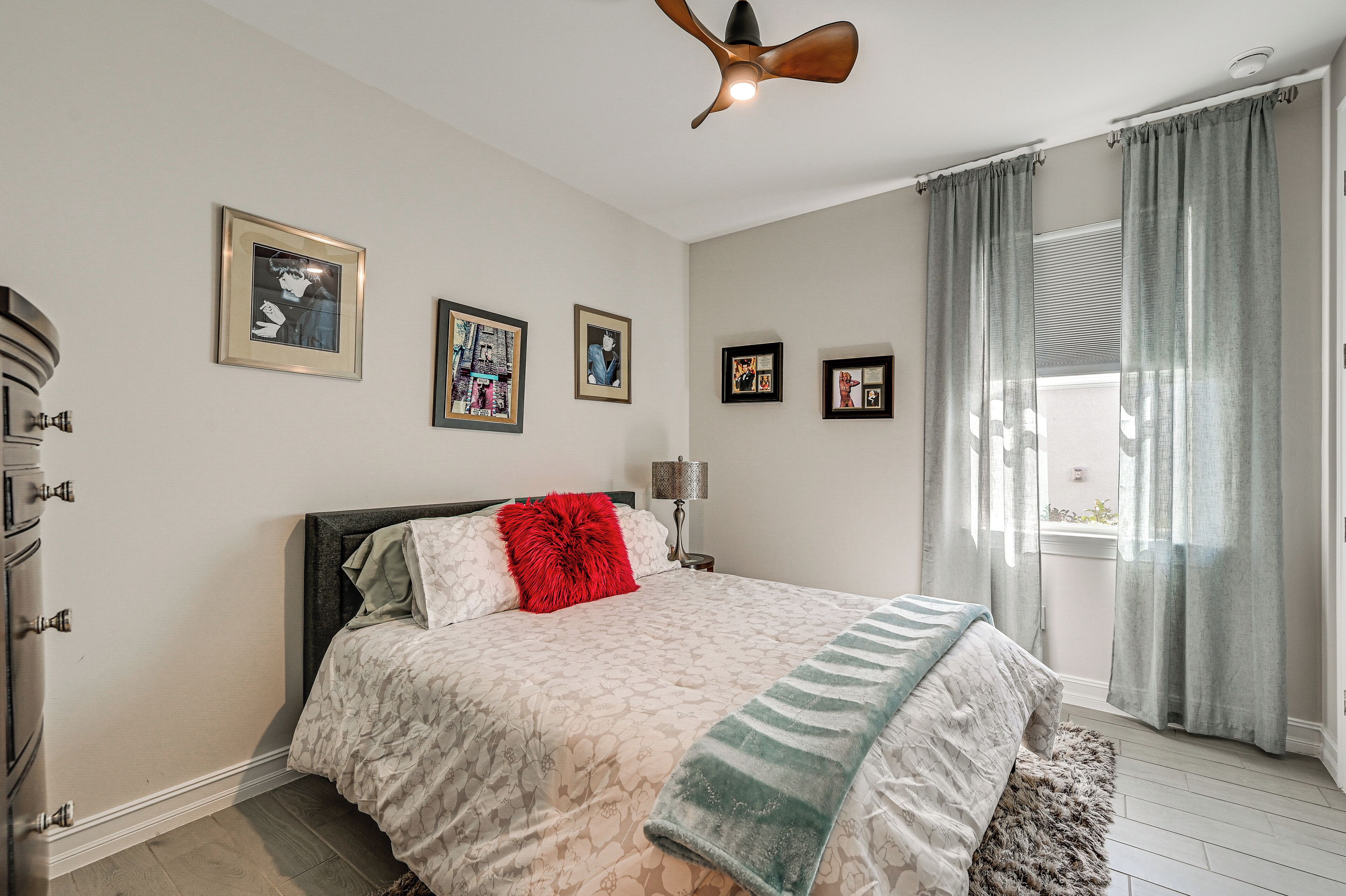 Ein Schlafzimmer mit Bett, Deckenventilator, Fenster mit Vorhängen und Wandkunst.