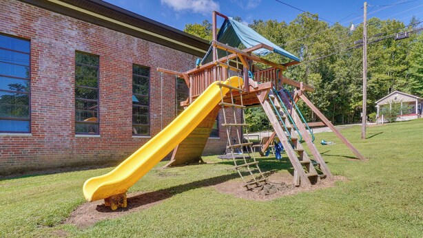 A playground with a yellow slide and a wooden climbing structure, set against a brick building with large windows. - ATV Trail Access: Mountain-view Briceville Studio! (Briceville)