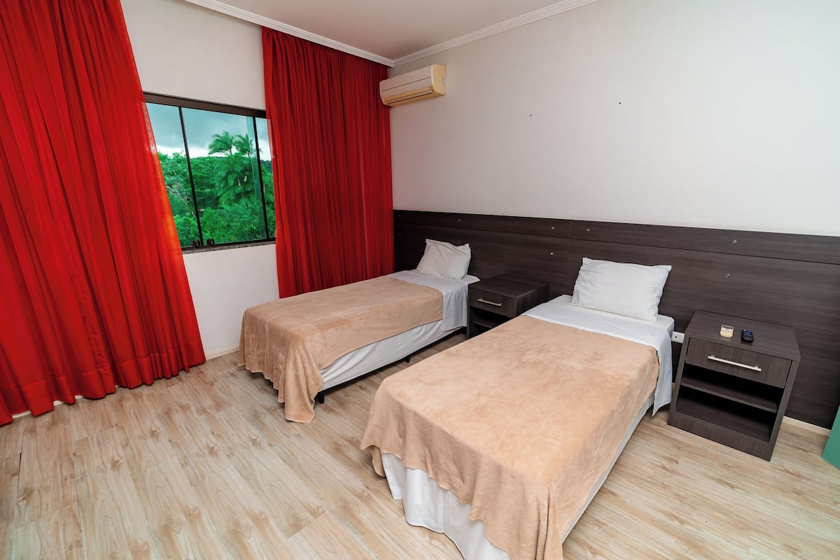 Ein Hotelzimmer mit zwei Betten, einem Nachttisch, einem Fenster mit roten Vorhängen und einer Klimaanlage.