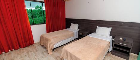 A hotel room with two beds, a nightstand, a window with red curtains, and an air conditioning unit.