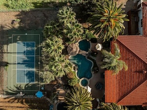 Vista aérea de una propiedad con cancha de tenis, piscina y palmeras a su alrededor.