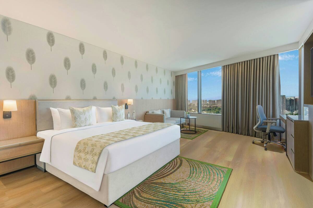 A hotel room with a large bed, a desk, a chair, a sofa, and a view of the city through large windows.