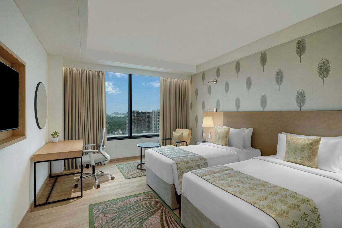 A hotel room with two beds, a desk, a chair, a TV, and a window with a city view.