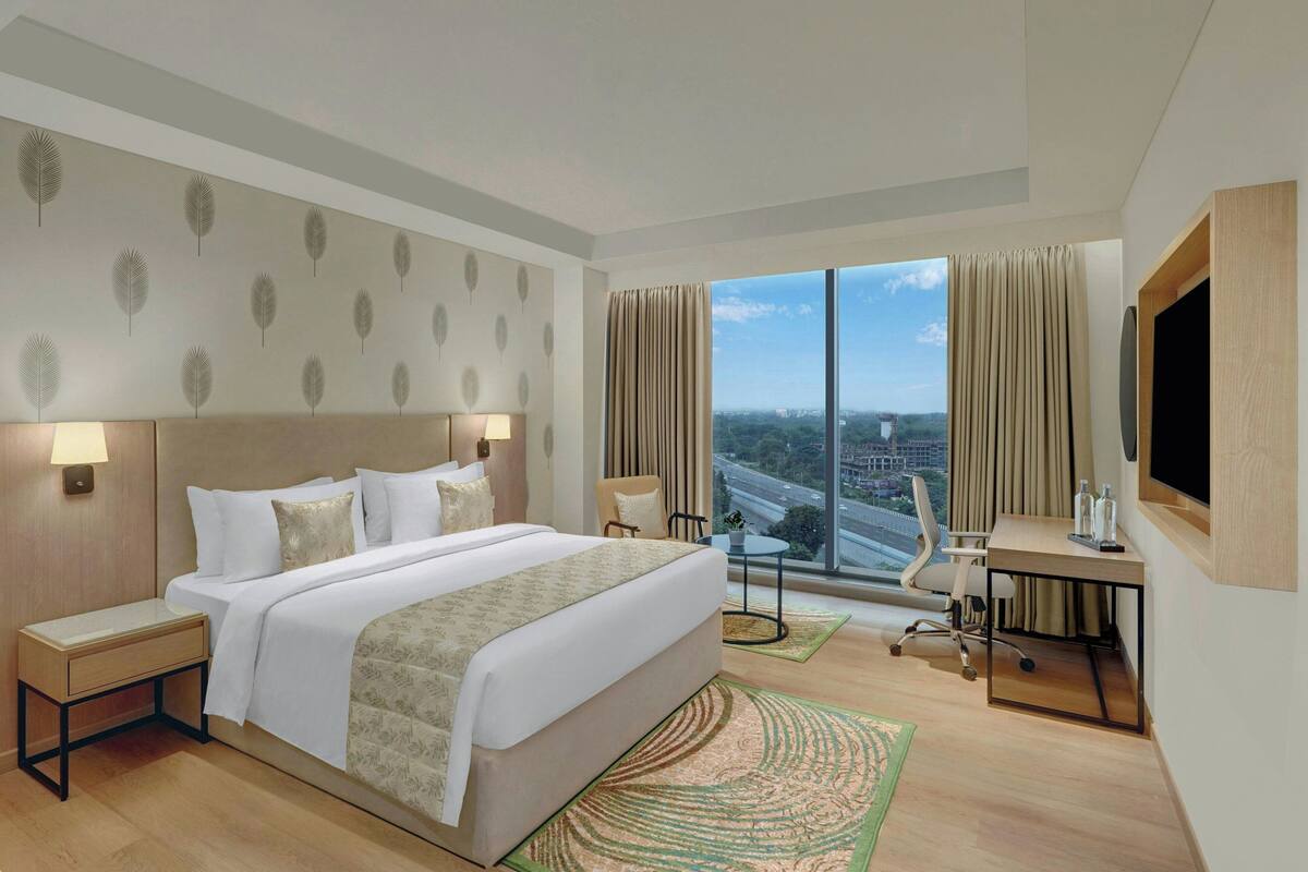 A modern hotel room with a large bed, a desk, a chair, and a view of the cityscape through large windows.