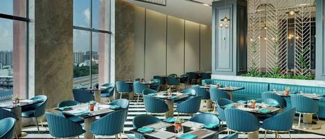 A modern restaurant with high ceilings, large windows, and a city view. Tables are set with blue chairs and gold accents.