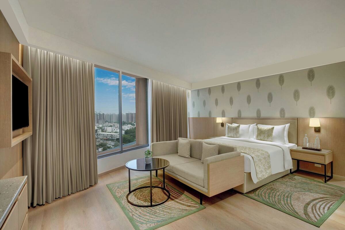 A hotel room with a bed, a sofa, a TV, a nightstand, and a view of the city.